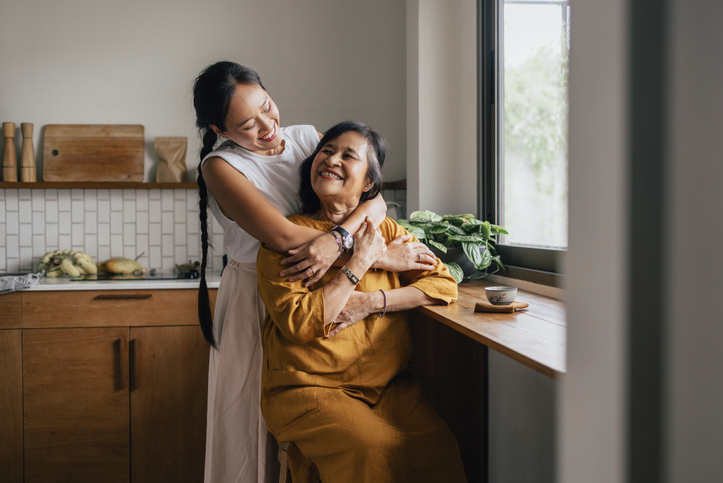 A smiling Asian woman enjoying drinking tea while her loving daughter is embracing her.
